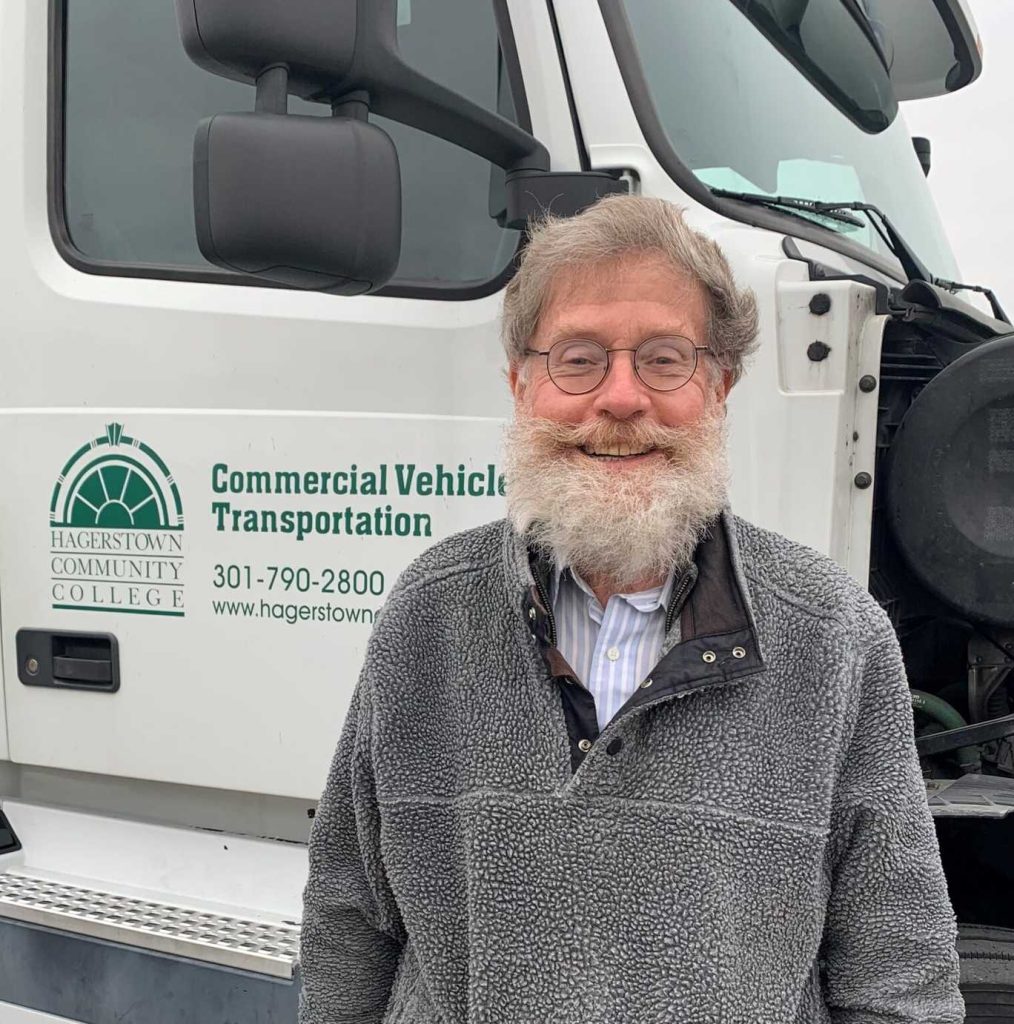 Older man with gray hair, beard, and glasses standing in front of a Commercial Vehicle Transportation truck.